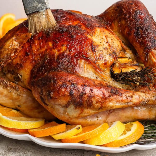 A fully cooked orange turkey on a platter with orange slices and being basted with a basting brush.