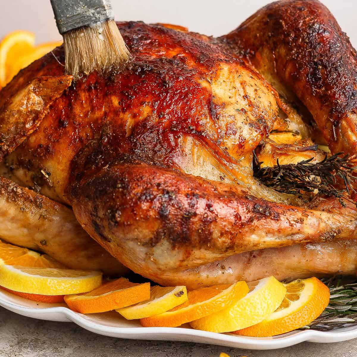 A fully cooked orange turkey on a platter with orange slices and being basted with a basting brush.