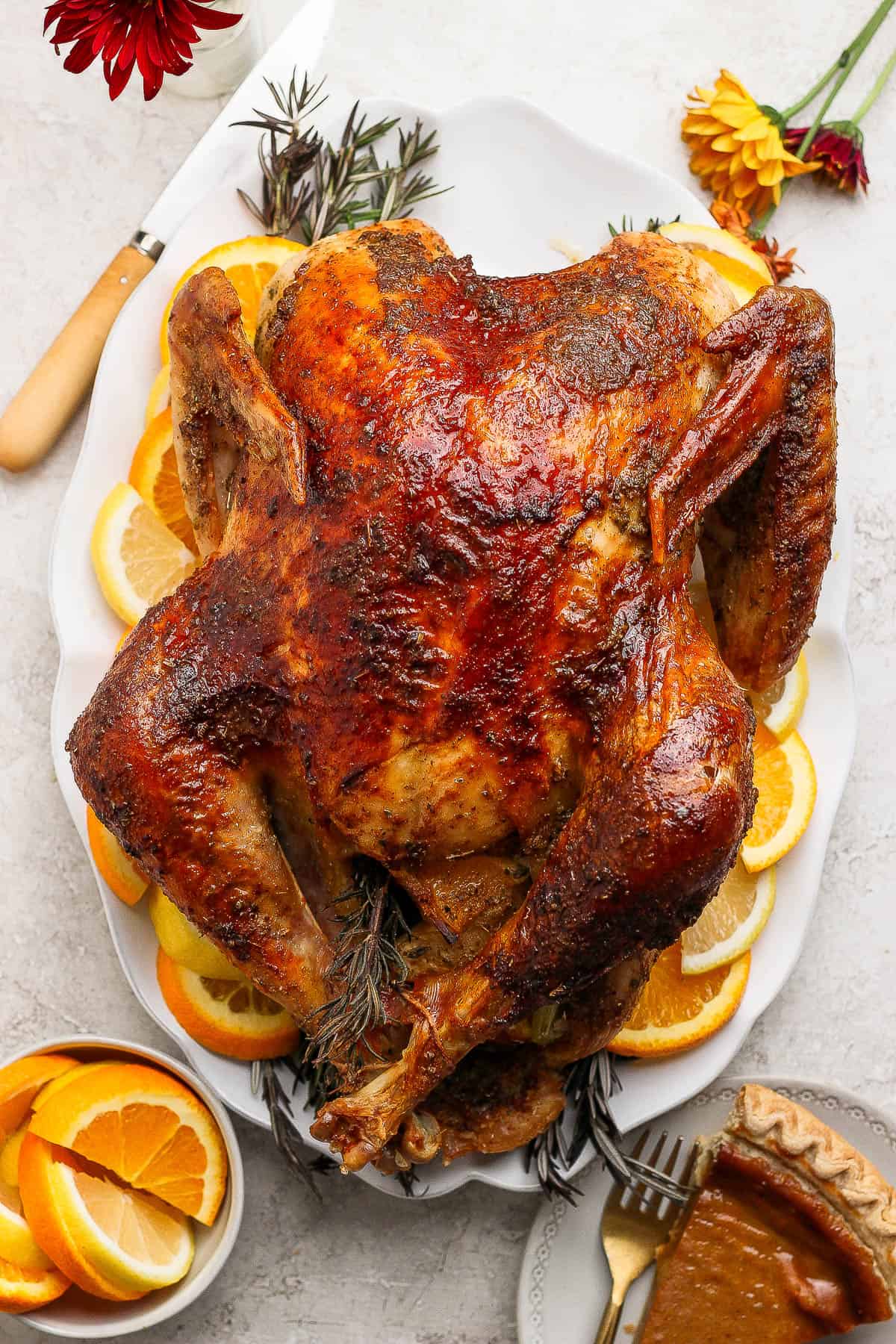 A fully cooked orange turkey on a white serving platter with orange slices and fresh rosemary next to a knife, bowl of orange slices, slice of pumpkin pie, and a couple daisies.