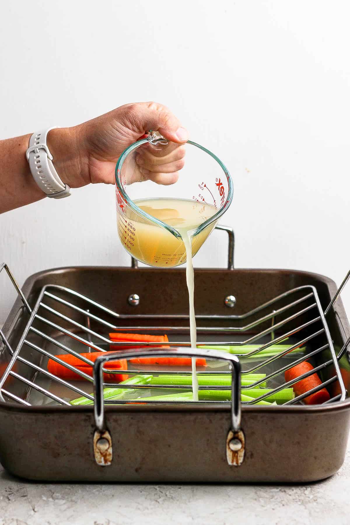 A glass measuring cup of chicken broth being poured into a roasting pan that has large chunks of carrots and celery.