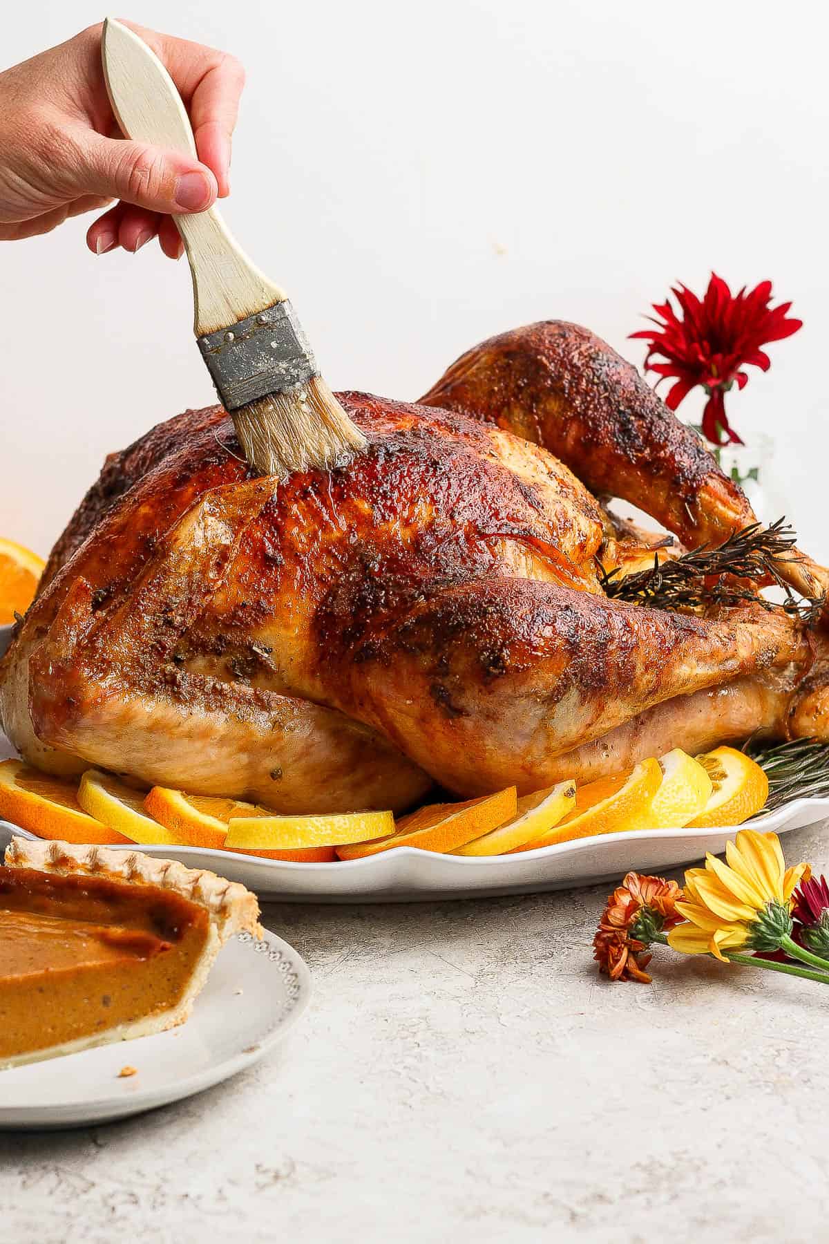 A fully cooked orange turkey on a platter with orange slices and fresh rosemary being basted with a basting brush.
