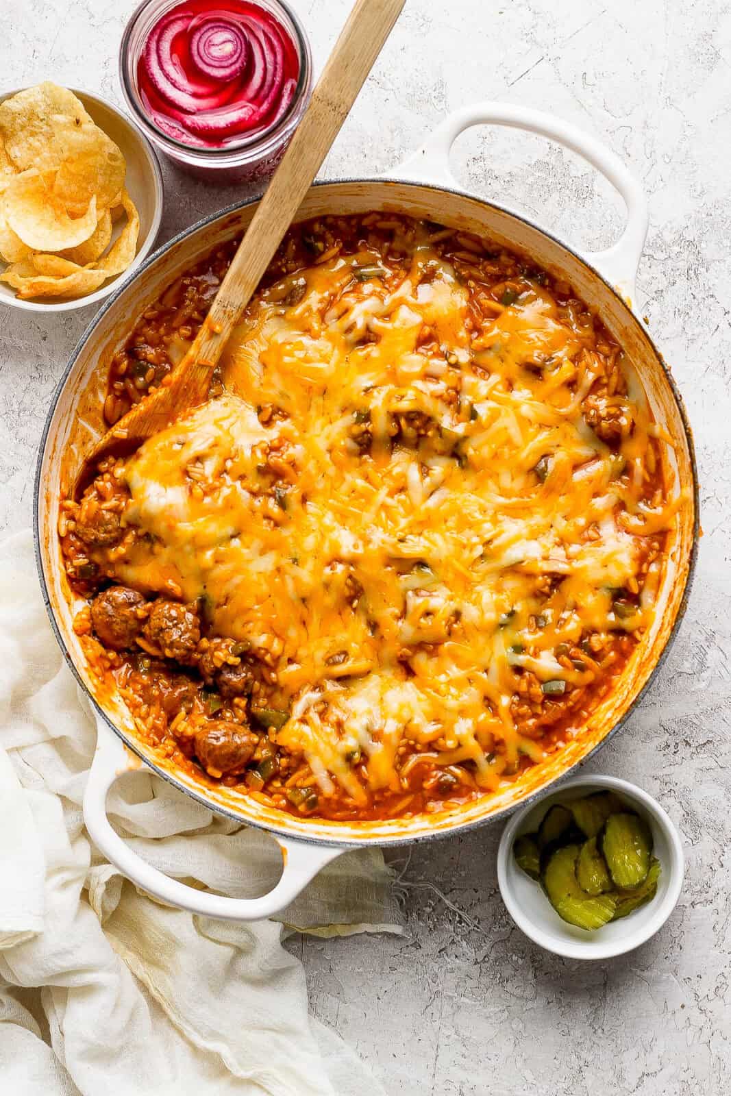 Fully cooked sloppy joe orzo with cheese on top and a wooden spoon in the side next to small bowls of chips, burger pickles, and pickled red onions.