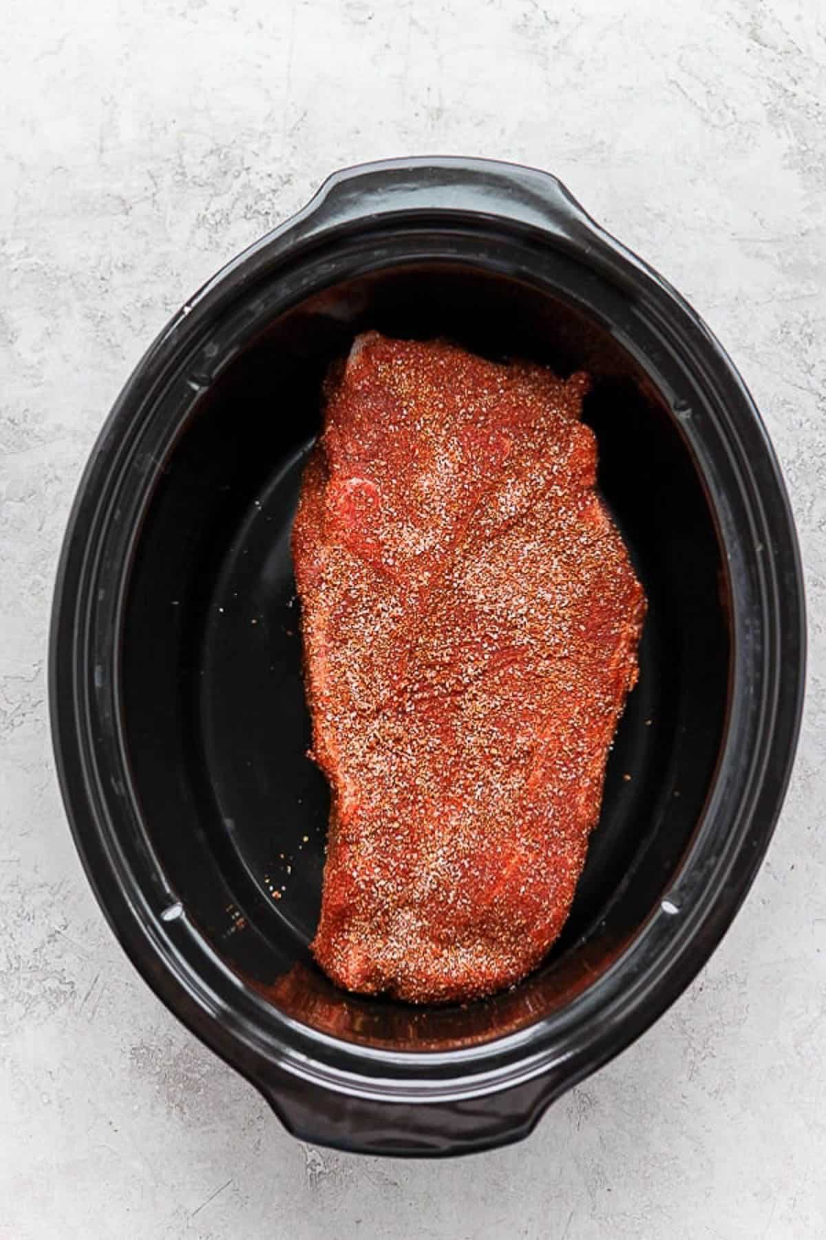 A bone-in pork shoulder covered in liquid smoke, yellow mustard, and dry rub in a slow cooker.