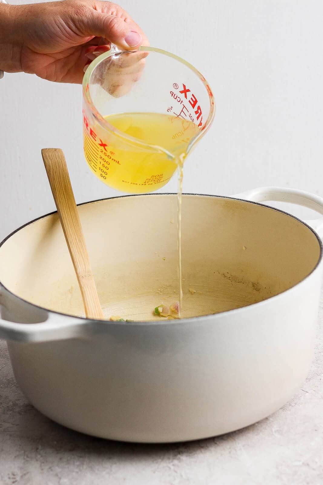 A measuring cup of chicken broth being poured in to a dutch oven with a wooden spoon in it.