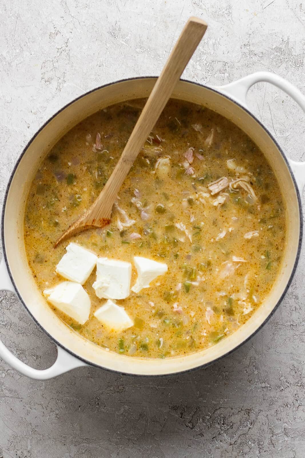 A pot of Mexican street corn white chicken chili with the cubes of cream cheese added and a wooden spoon sticking out the side.