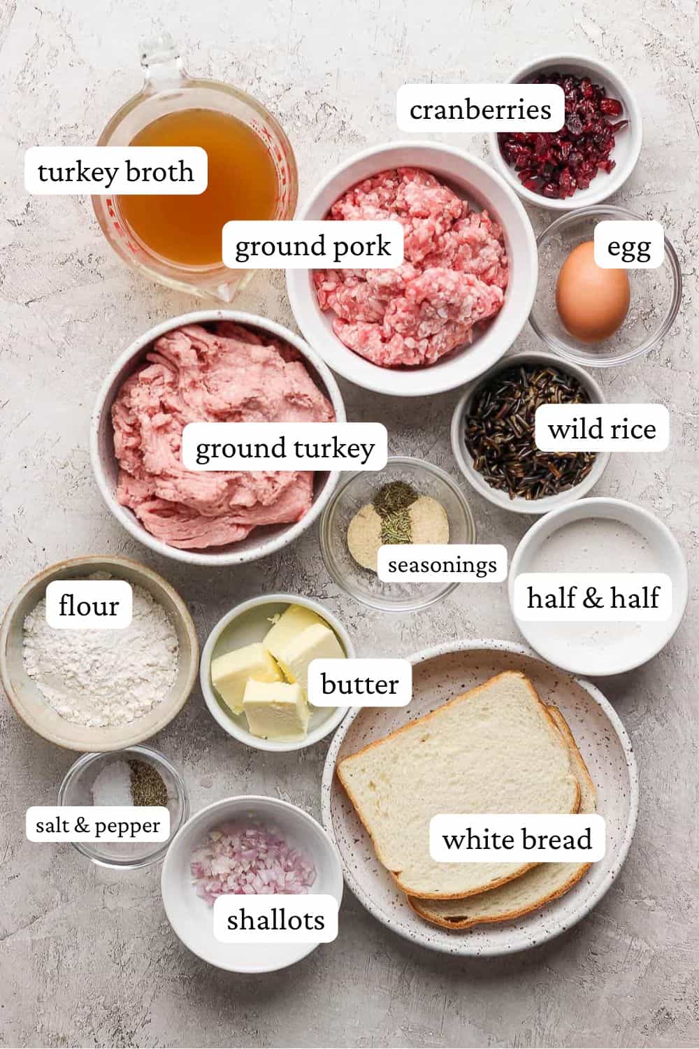 Thanksgiving meatball ingredients in individual bowls; turkey broth, dried cranberries, ground pork, egg, ground turkey, wild rice, seasonings, half & half, flour, butter, 2 slices of white bread, shallots, and salt and pepper.