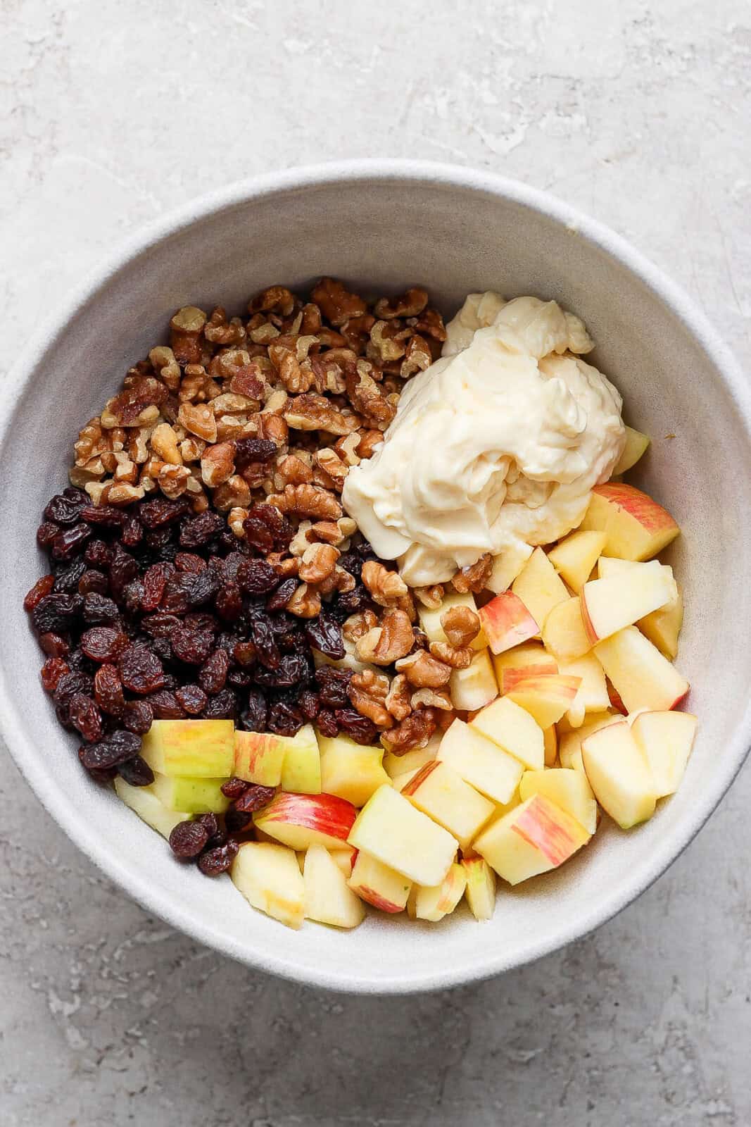 Apples, walnuts, raisins, and mayo unmixed in a bowl to make apple and walnut salad.