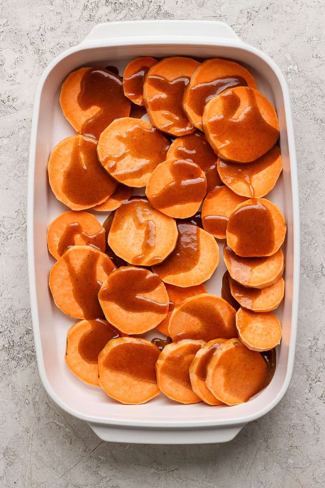 A layer of peeled and sliced sweet potatoes covering the bottom of a casserole dish and drizzled with half of the butter sugar mixture.