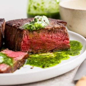 A cooked filet mignon on a white plate with some chimichurri and herbed butter on top.