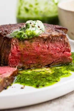A cooked filet mignon on a white plate with some chimichurri and herbed butter on top.