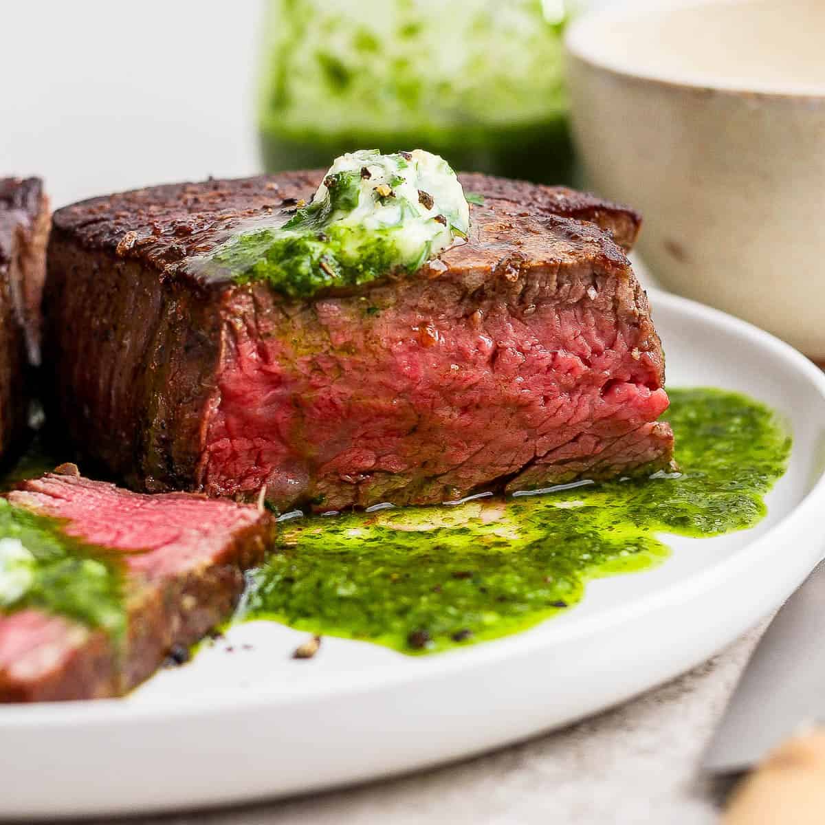A cooked filet mignon on a white plate with some chimichurri and herbed butter on top.