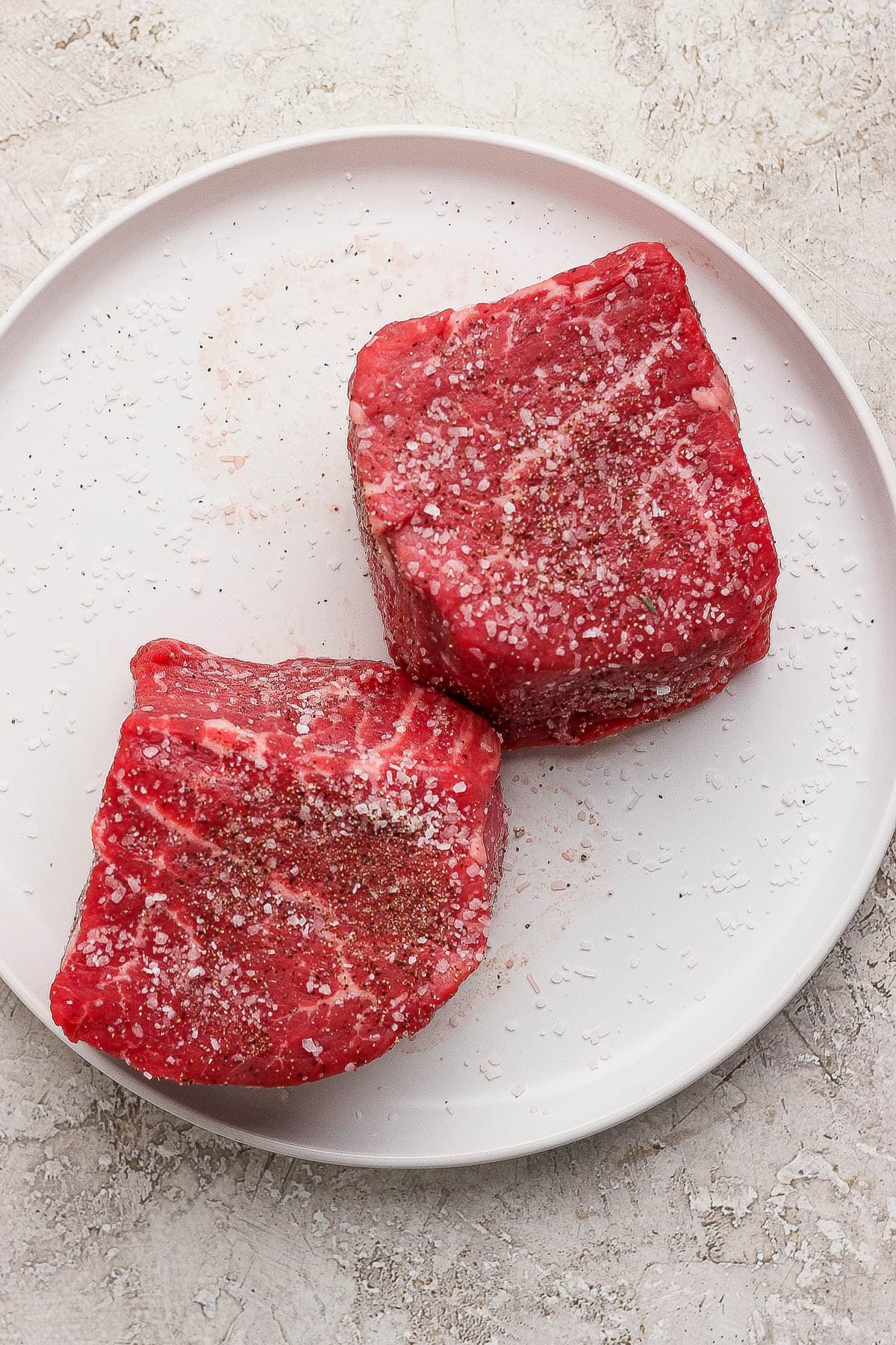Two filet mignon steaks seasoned with salt and pepper on a white plate.