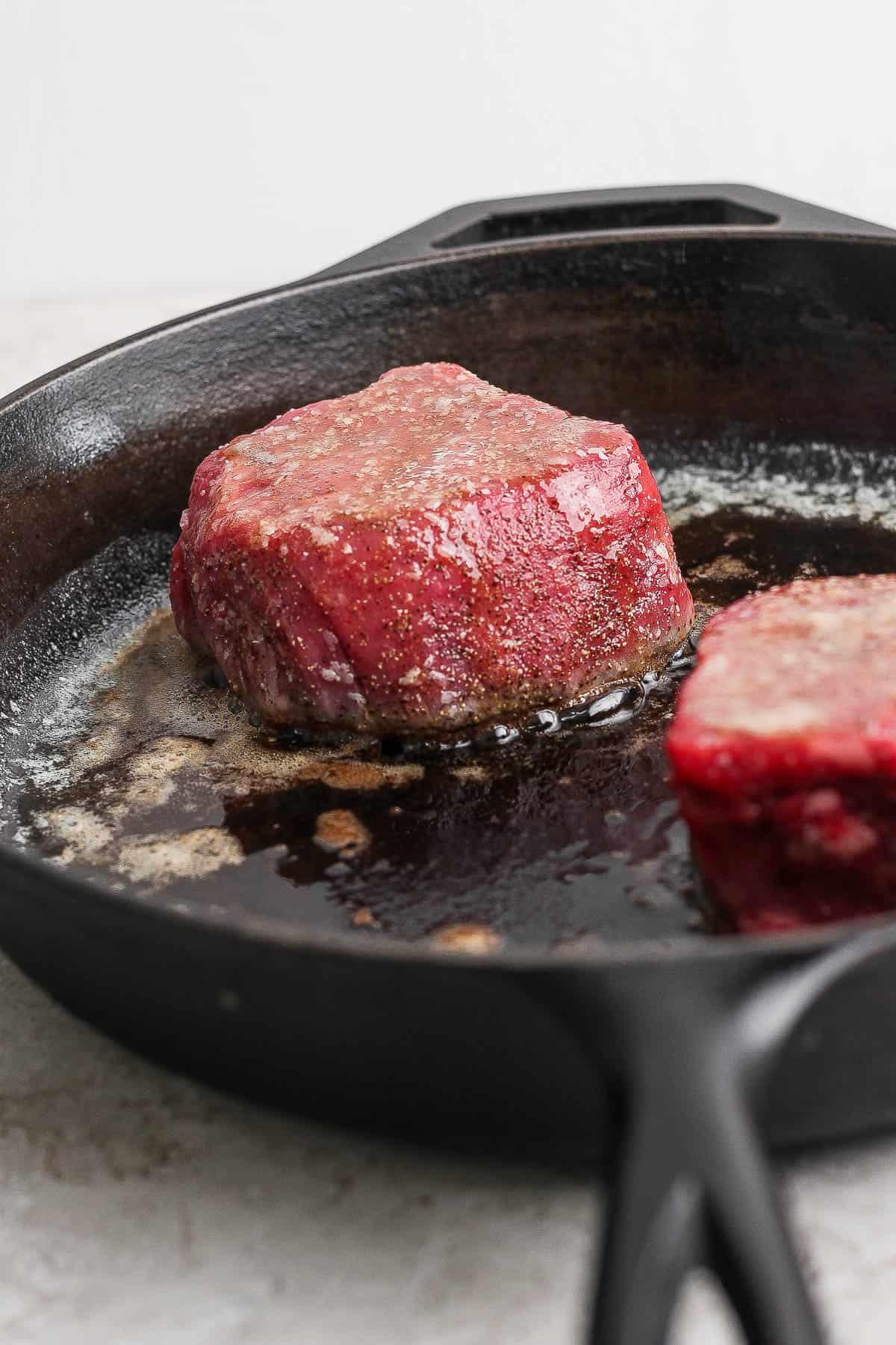 Two filet mignon steaks searing on one side in a cast iron skillet with melted butter.