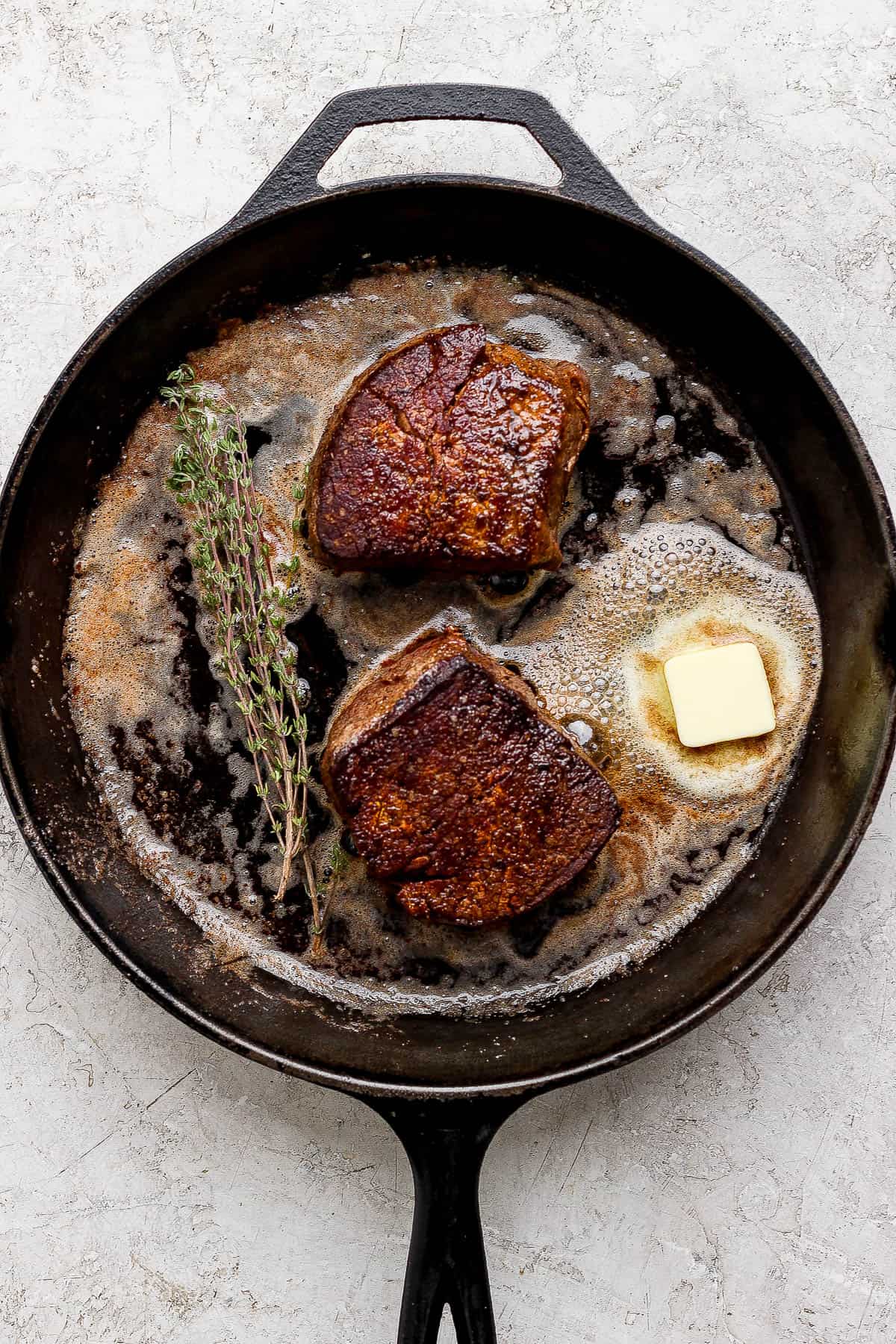 Two filet mignon steaks cooking in a cast iron skillet with a pad of butter and fresh thyme.