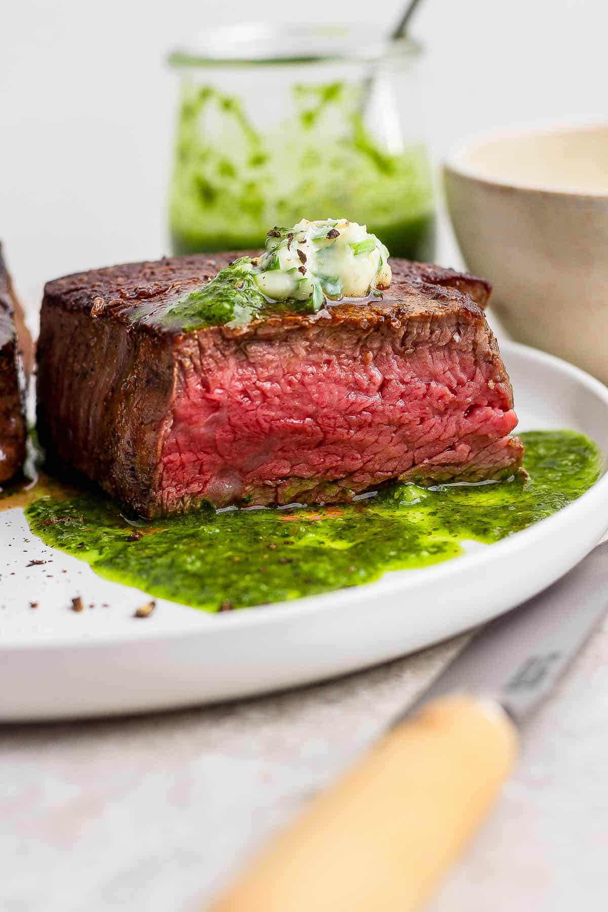A fully cooked cast iron skillet filet mignon after being cut in half on a white plate with chimichurri sauce and some garlic herb butter.