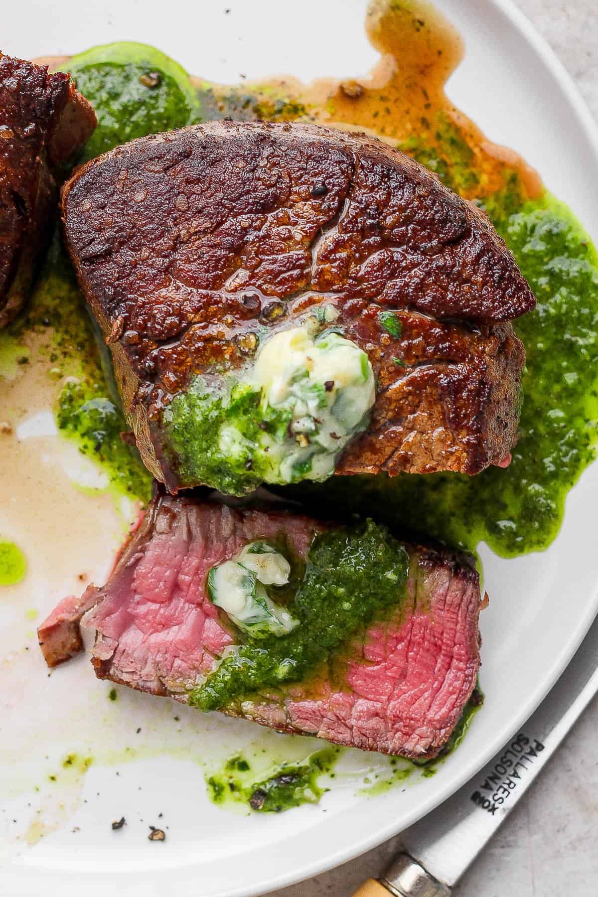 A cast iron skillet filet mignon on a white plate with chimichurri sauce and some garlic herb butter with a piece sliced off.
