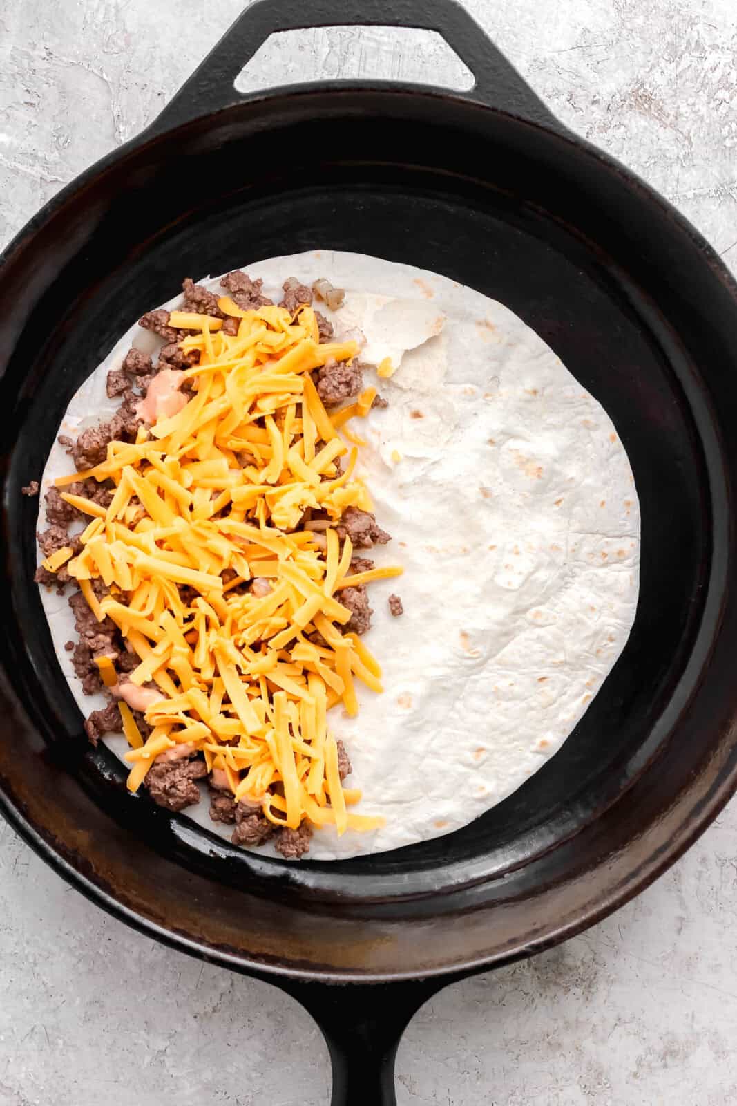 A tortilla in a cast iron skillet with meat mixture, burger sauce, and shredded cheese on one half to create a cheeseburger quesadilla.