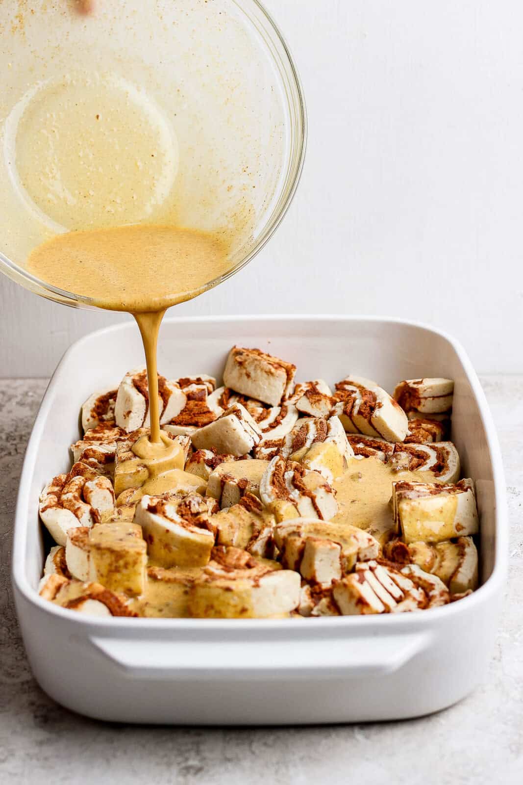 Quartered cinnamon rolls in a 9x13 baking dish with egg mixture being poured over the top.