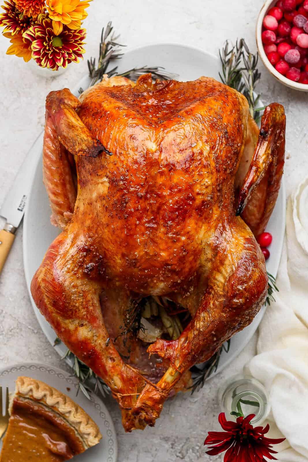 A cooked whole grilled turkey on a platter with cranberries and pumpkin pie around it.