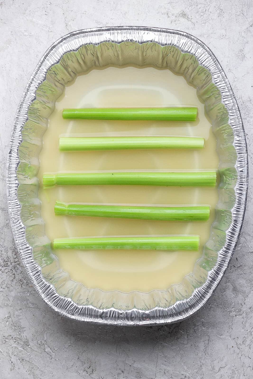 An aluminum pan with broth inside and celery sticks lined up to act like a roasting rack inside.
