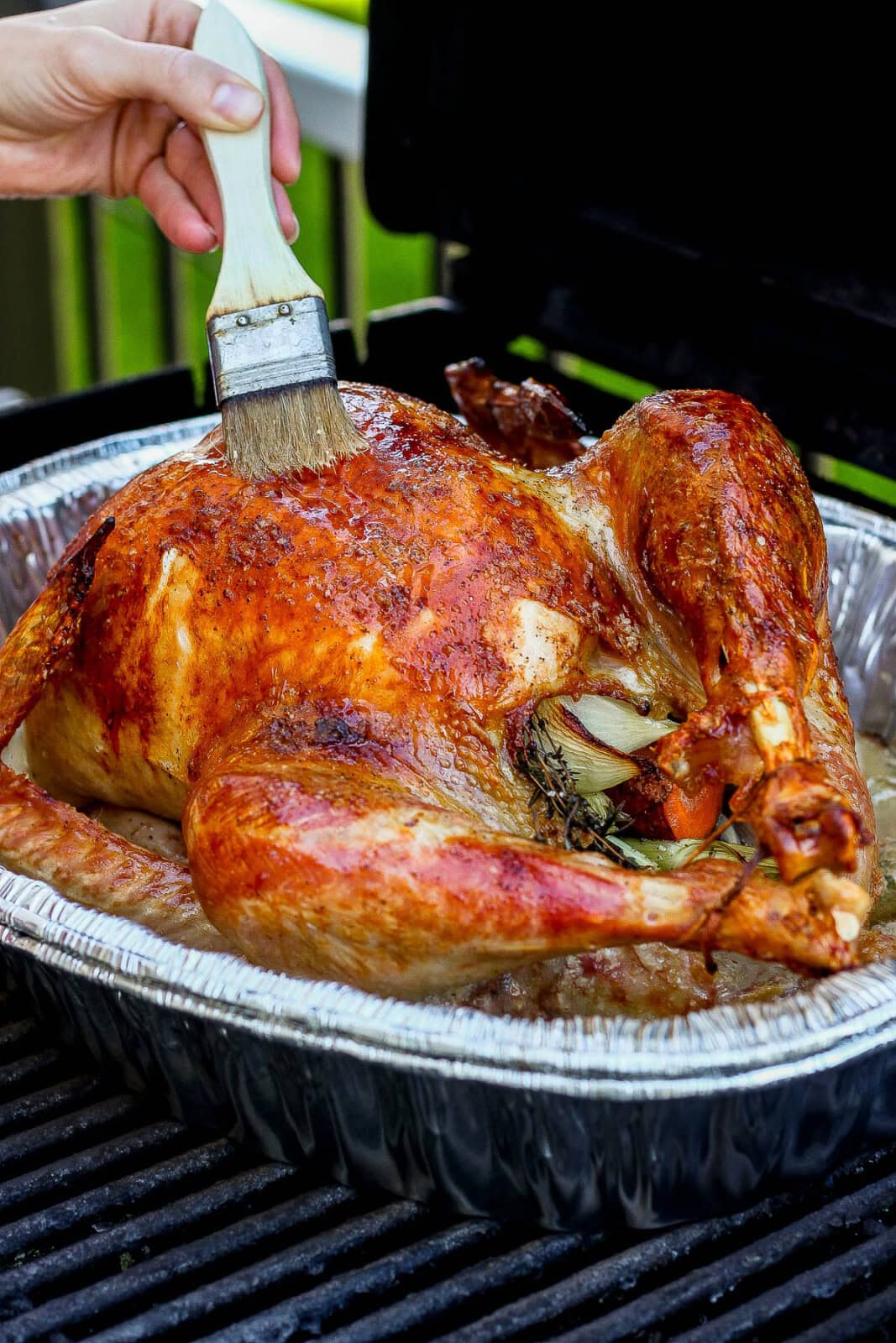 A cooked grilled turkey inside a roasting rack, still on the grill, with someone basting it with a brush.