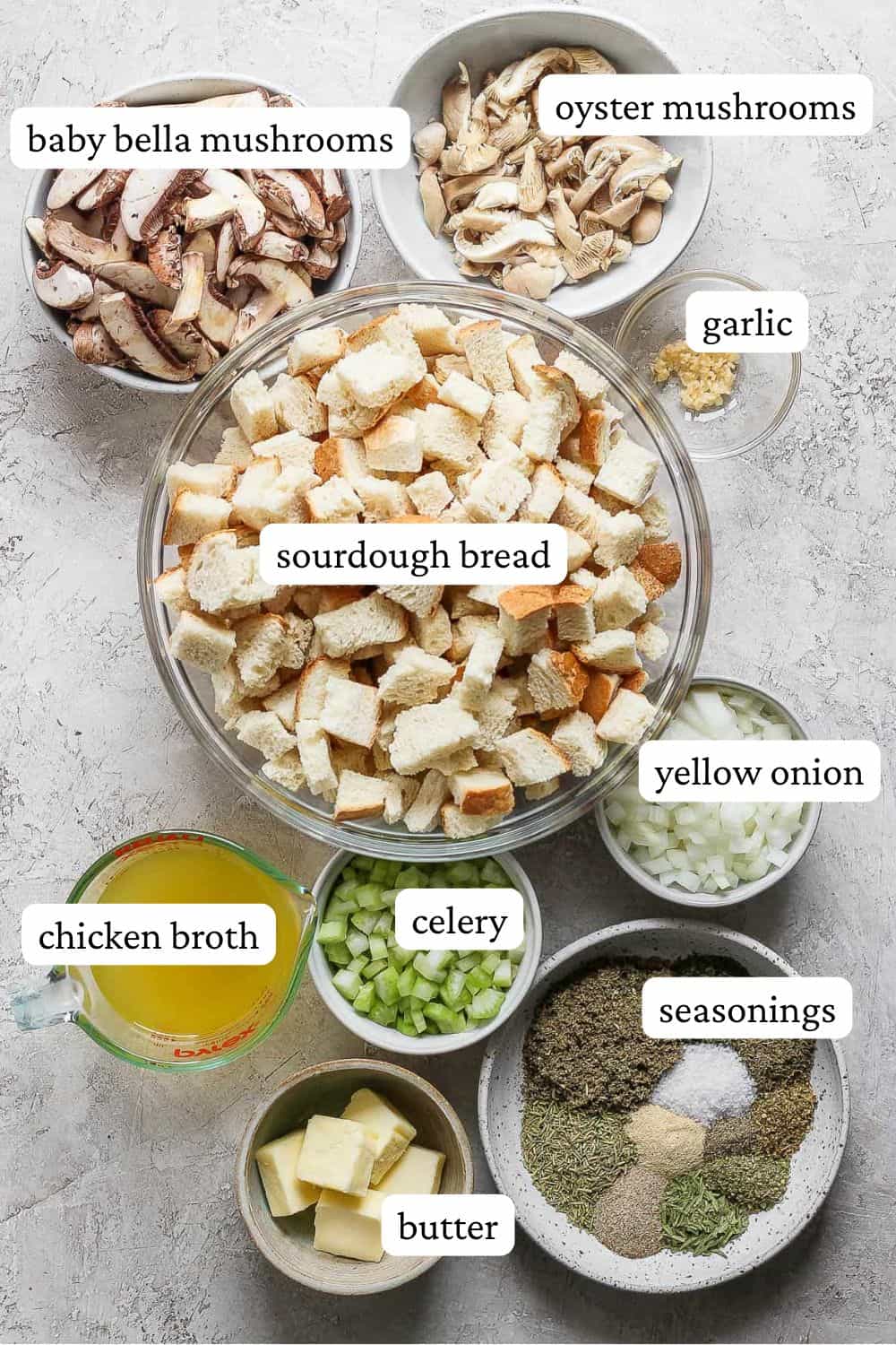 Ingredients for crock pot stuffing in individual bowls; baby bella mushrooms, oyster mushrooms, garlic, sourdough bread, yellow onion, chicken broth, celery, seasonings, and butter.