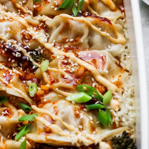 A casserole dish filled with a dumpling bake with rice, broccolini, dumplings (or potstickers), spicy mayo, sauce, green onions and sesame seeds.