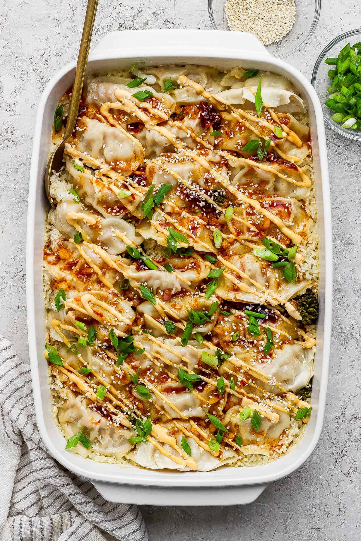 A fully cooked dumpling bake in a white baking dish with a gold spoon sticking out the side and two bowls on the side with sesame seeds and green onions.