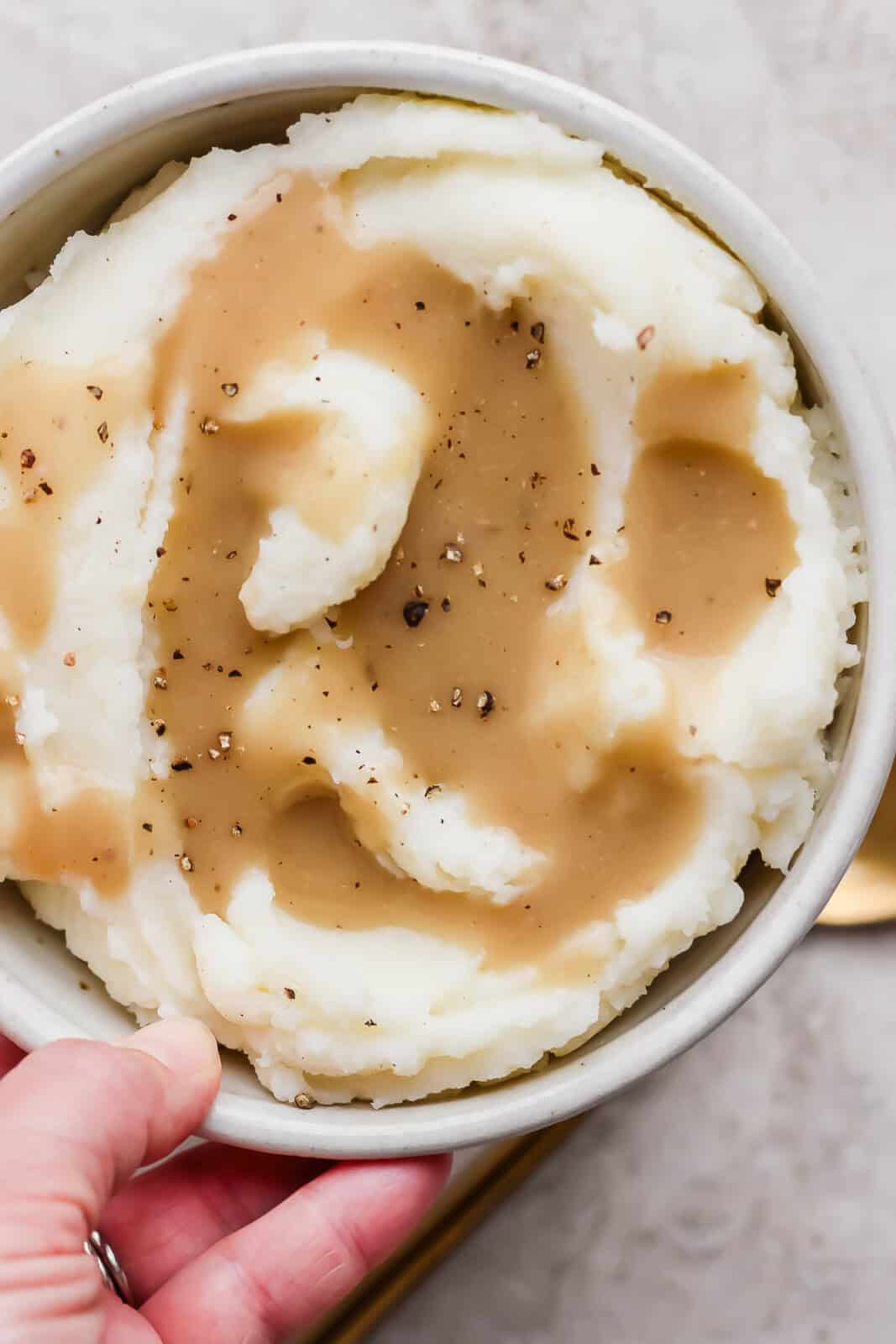 Someone holding a bowl of mashed potatoes with gluten and dairy free gravy on top with fresh cracked pepper.