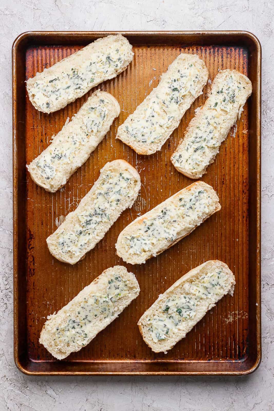 Eight halves of hot dog buns opened on a baking sheet and spread with herbed butter.