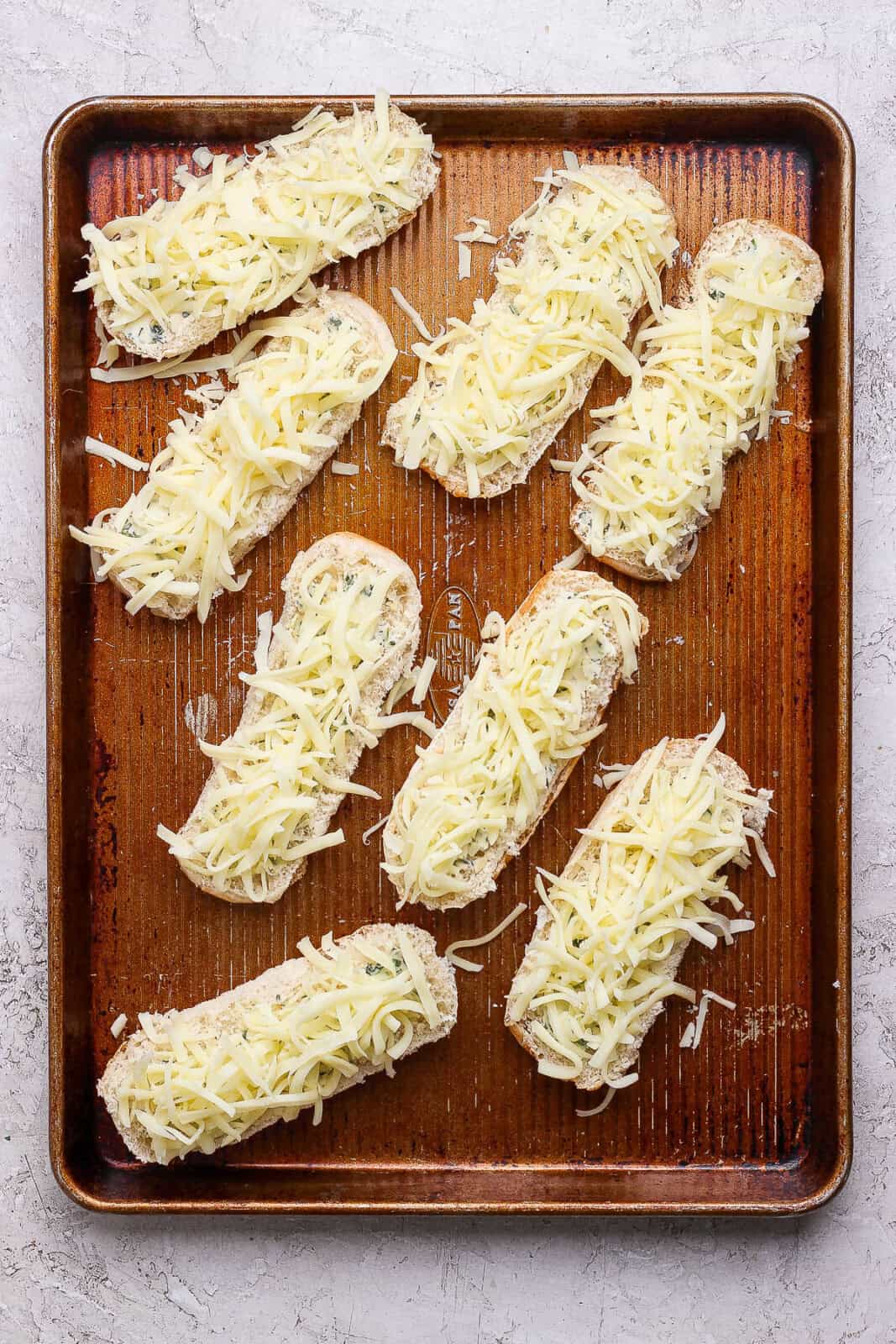 8 halved and opened hotdog buns on a baking sheet topped with herbed cheese and shredded mozzarella cheese.