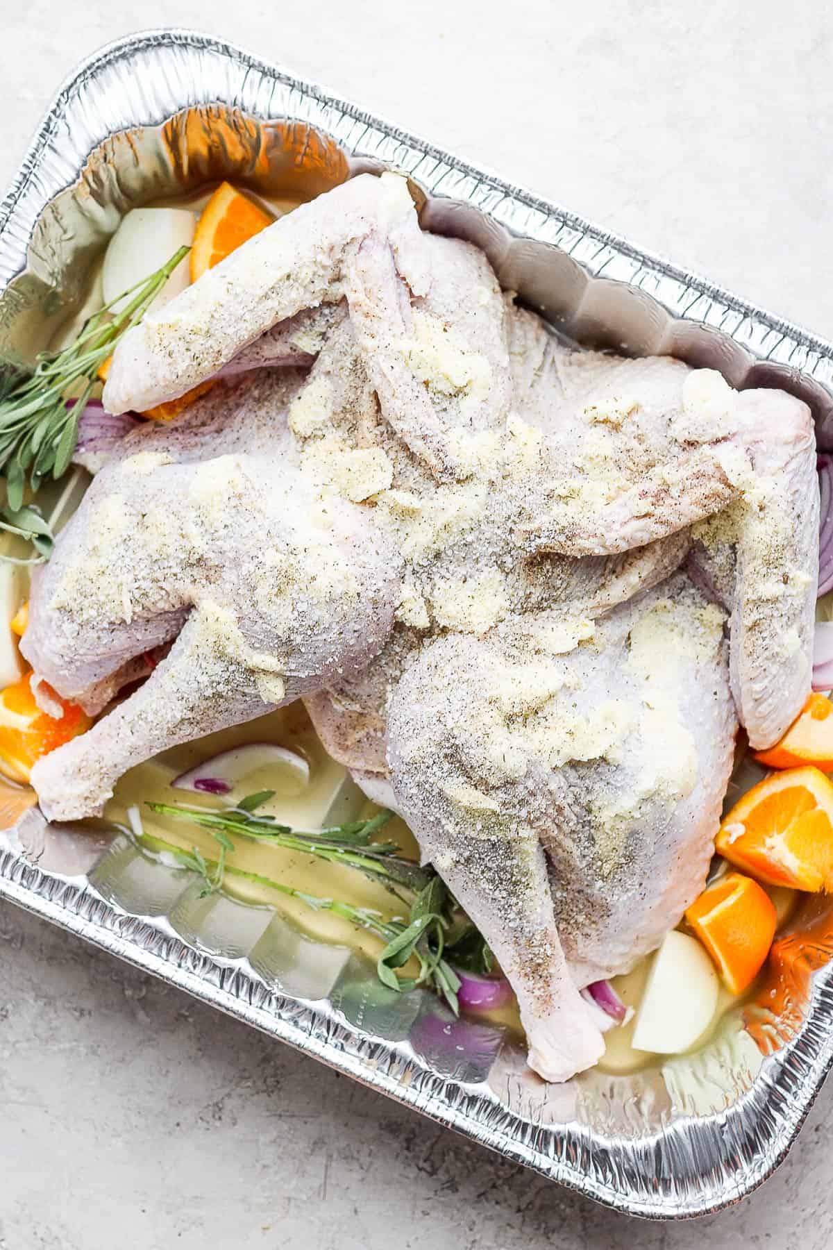 A spatchcock turkey rubbed with butter and seasoned sitting in a prepared aluminum roasting pan.