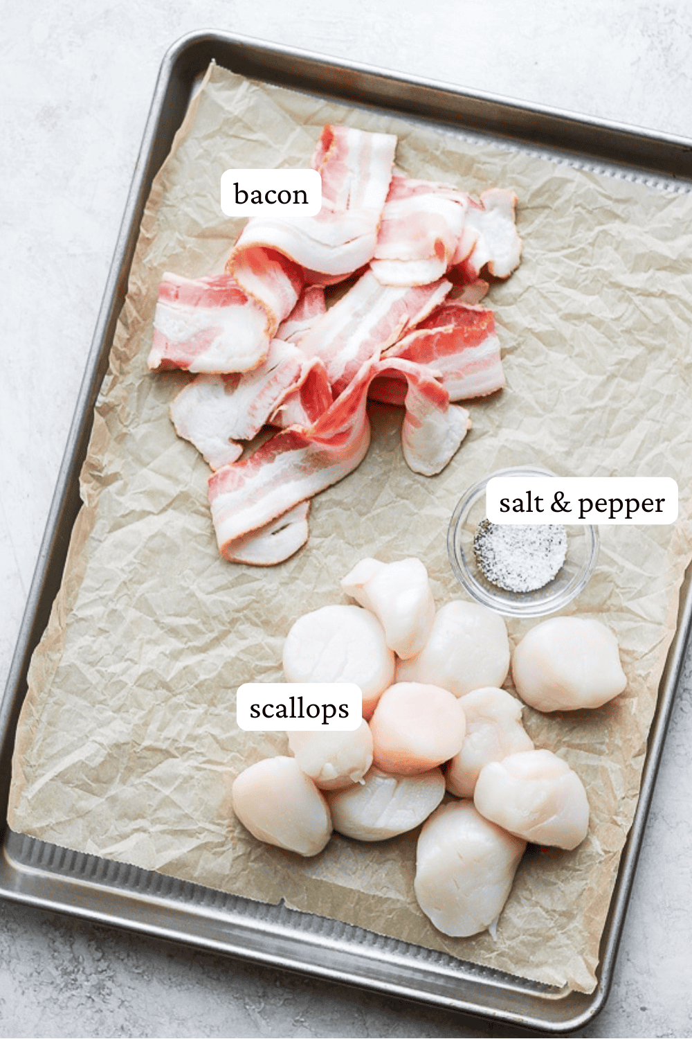 Bacon strips, fresh scallops, and a small bowl of salt & pepper on a parchment lined baking sheet.