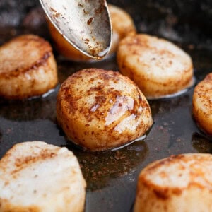 Several scallops cooking in browned butter in a skillet with a spoon drizzling butter over one of them.