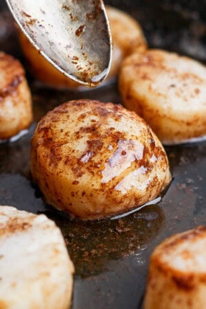 Several scallops cooking in browned butter in a skillet with a spoon drizzling butter over one of them.