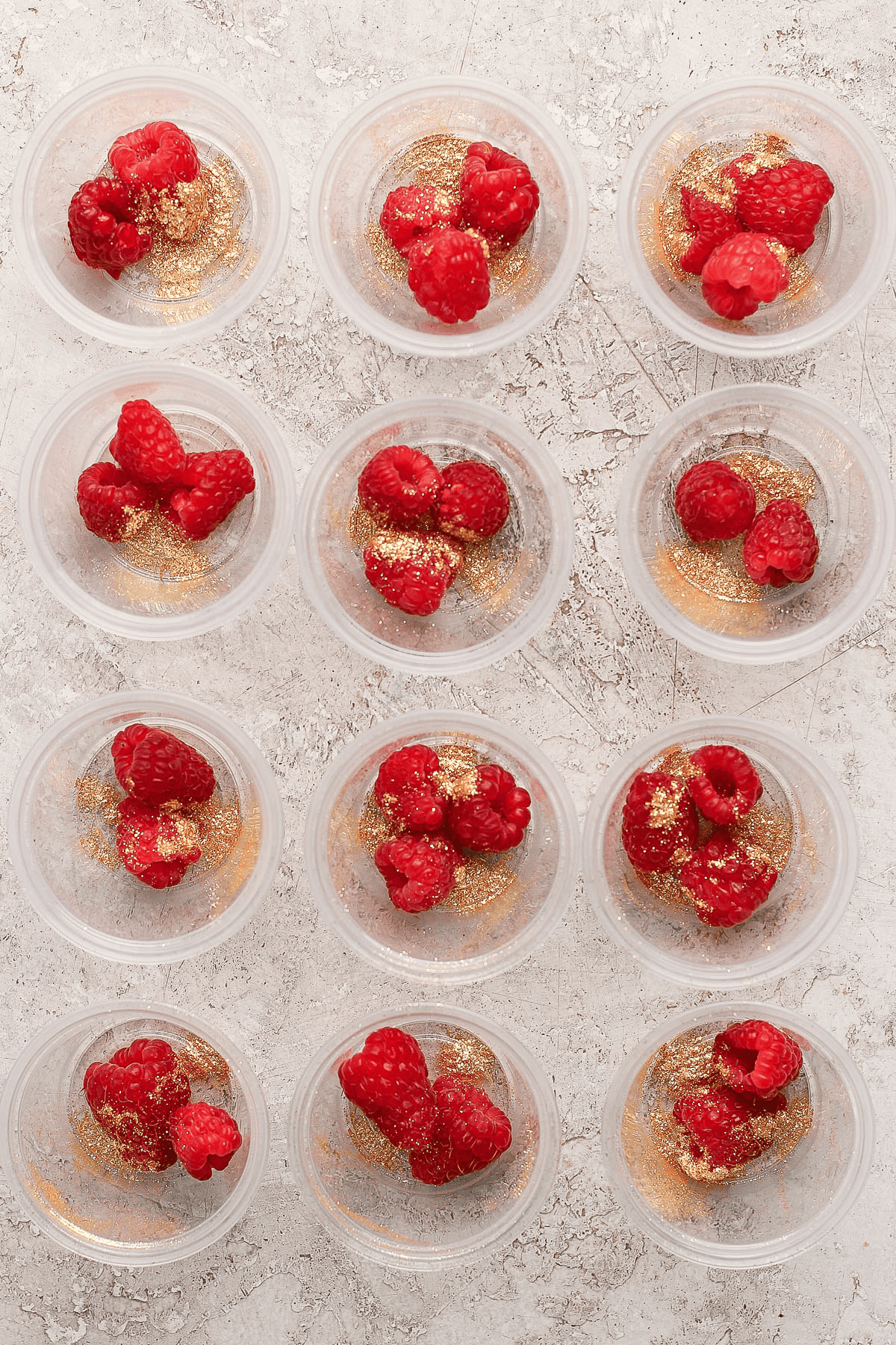 Twelve plastic portion cups with fresh raspberries and edible gold glitter in them.