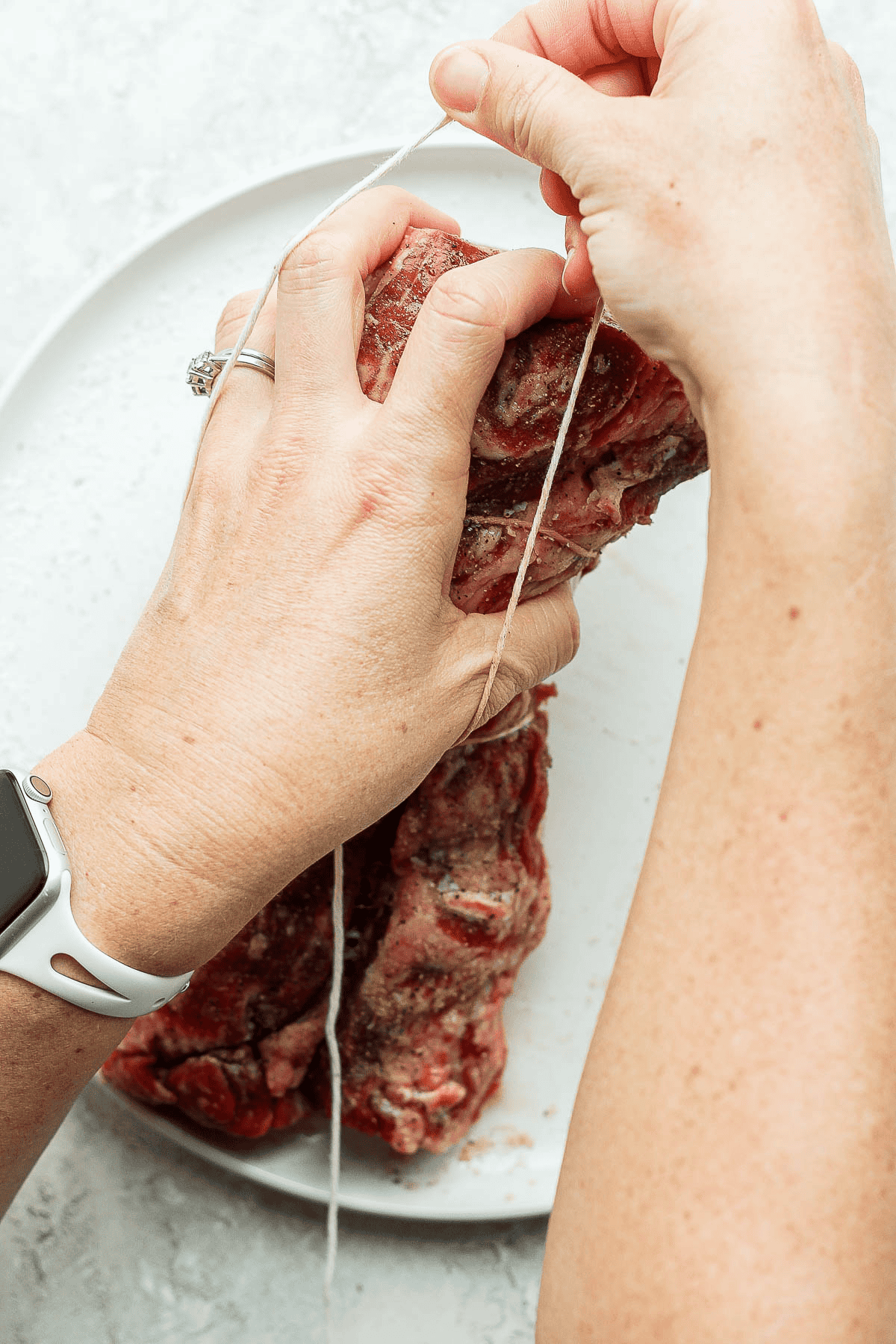 One hand holding the beef tenderloin and another hand pulling twine off the hand and over the tenderloin.