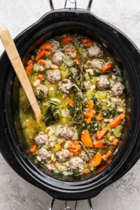 Italian wedding soup in a slow cooker with a wooden spoon sticking out the side.