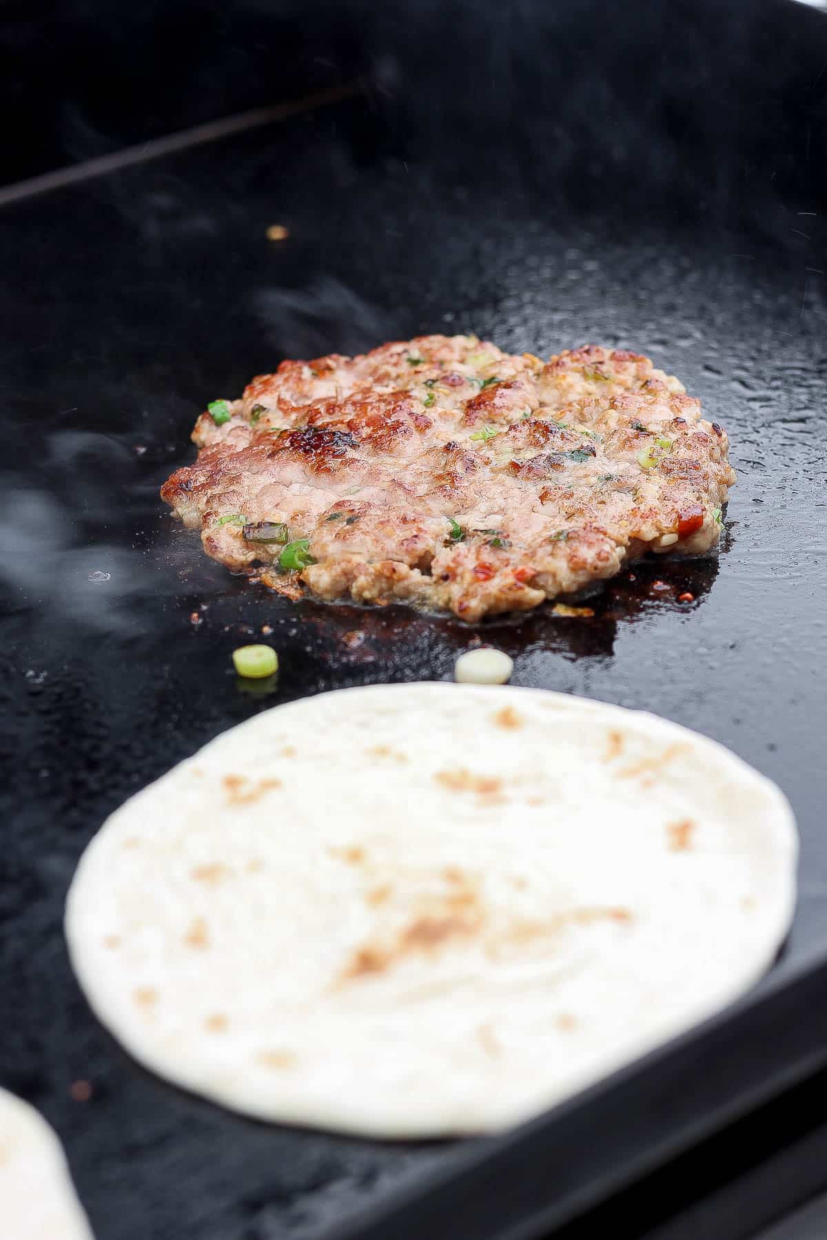 A pork patty on a hot griddle next to a street taco tortilla.