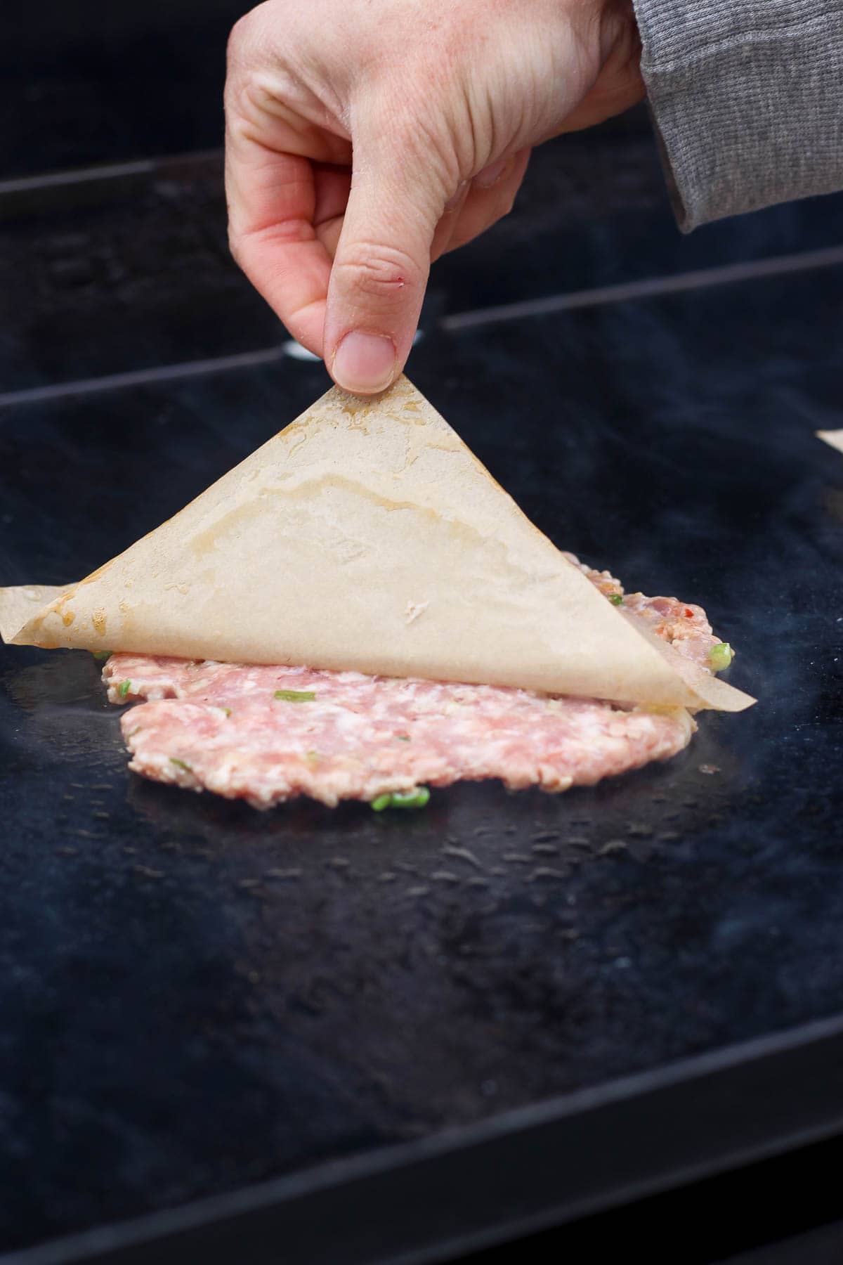 A smashed pork patty for smash dumpling tacos on a hot griddle with a hand peeling off the piece of parchment paper.