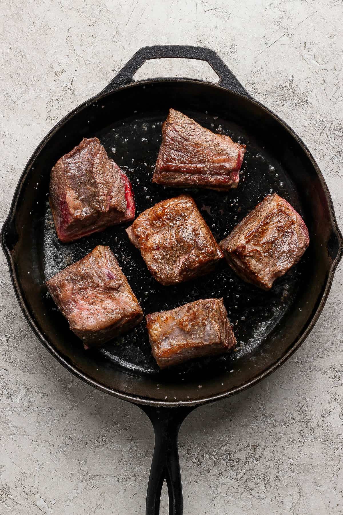 Six short ribs being braised in a cast iron skillet for short rib tacos.