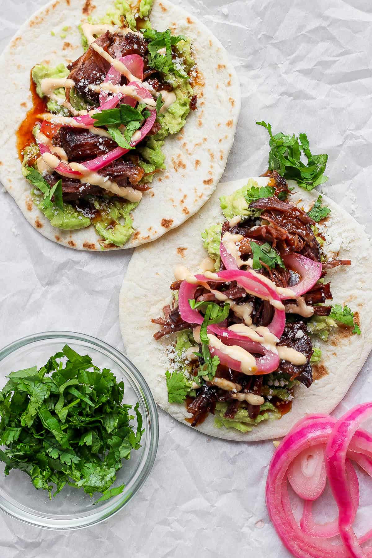 Two short rib tacos assembled with toppings of spicy mayo, pickled onions, cilantro, and smashed avocado.