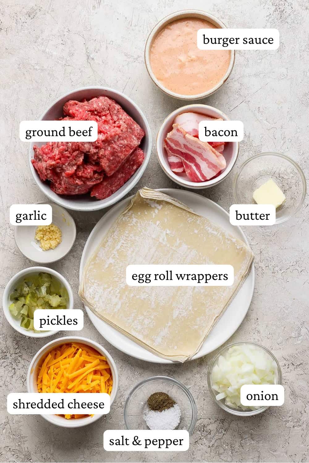 Cheeseburger egg roll ingredients in individual bowls; burger sauce, ground beef, bacon, butter, egg roll wrappers, garlic, pickles, shredded cheddar cheese, salt and pepper, and diced onion.