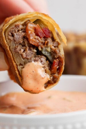 A hand holding a cheeseburger egg roll over a bowl of burger sauce.