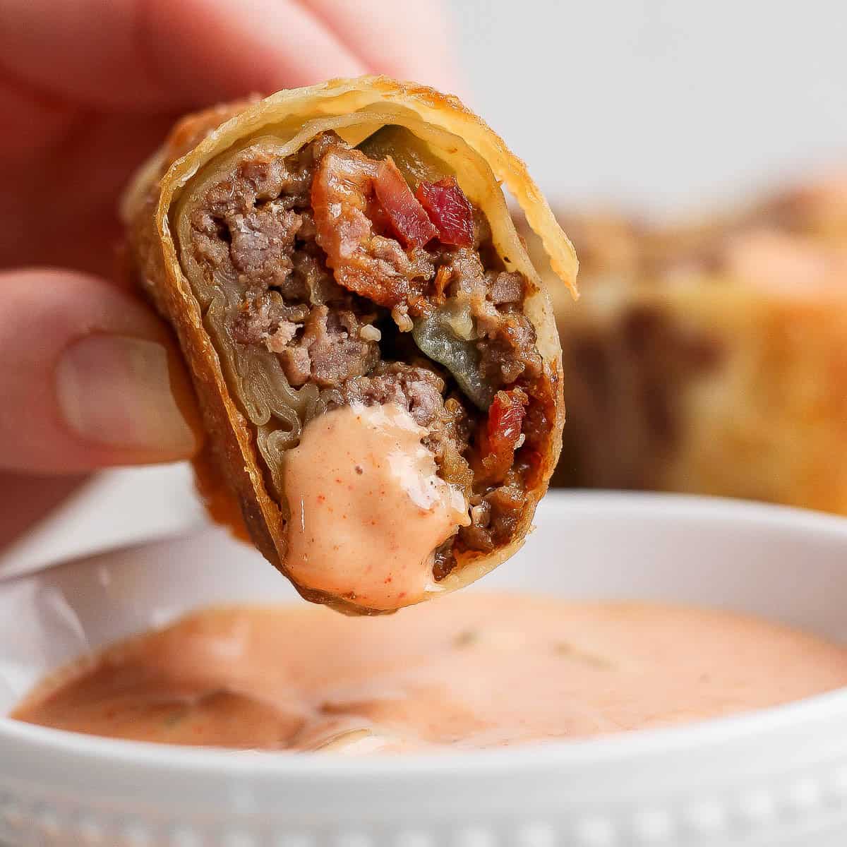 A hand holding a cut cheeseburger egg roll over a bowl of burger sauce.