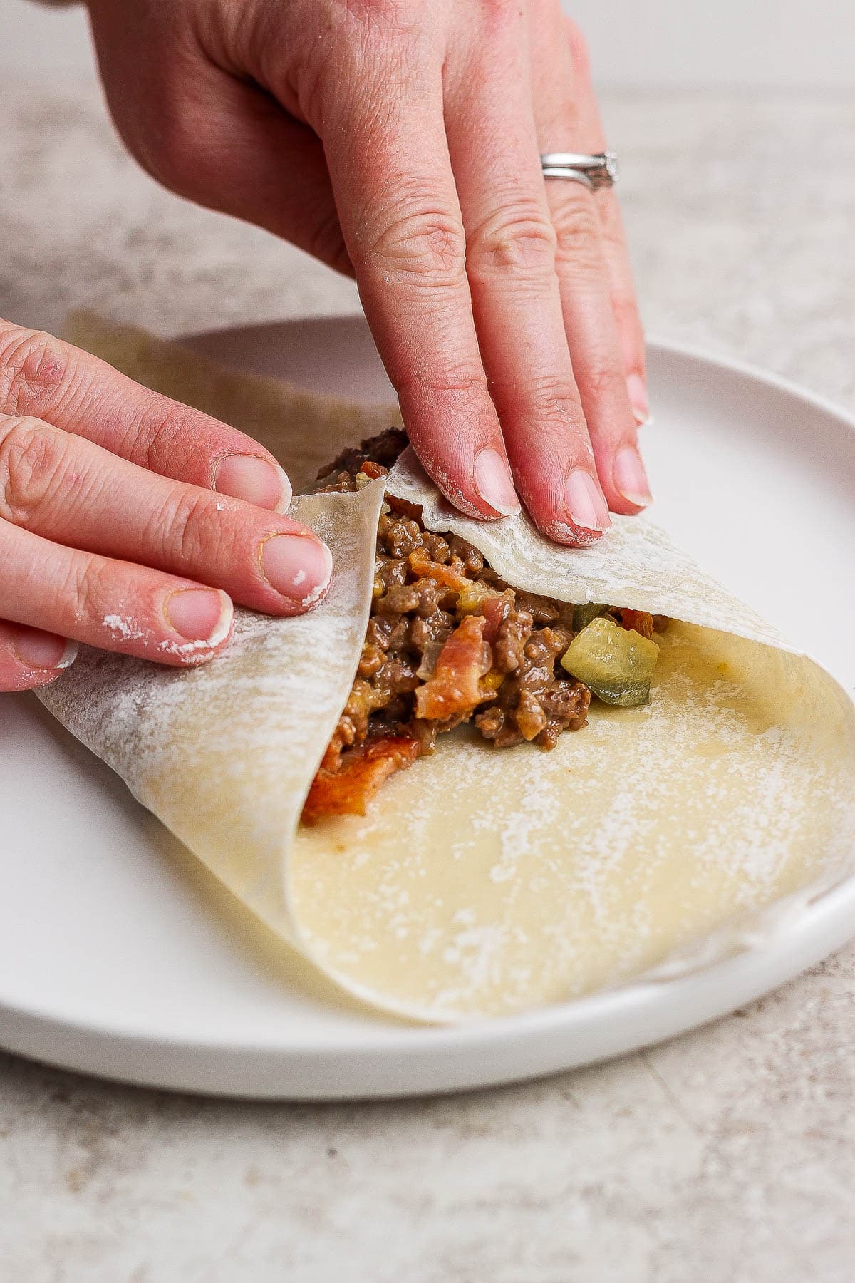 Hands folding the egg roll wrapper corners towards the center to make cheeseburger egg rolls.