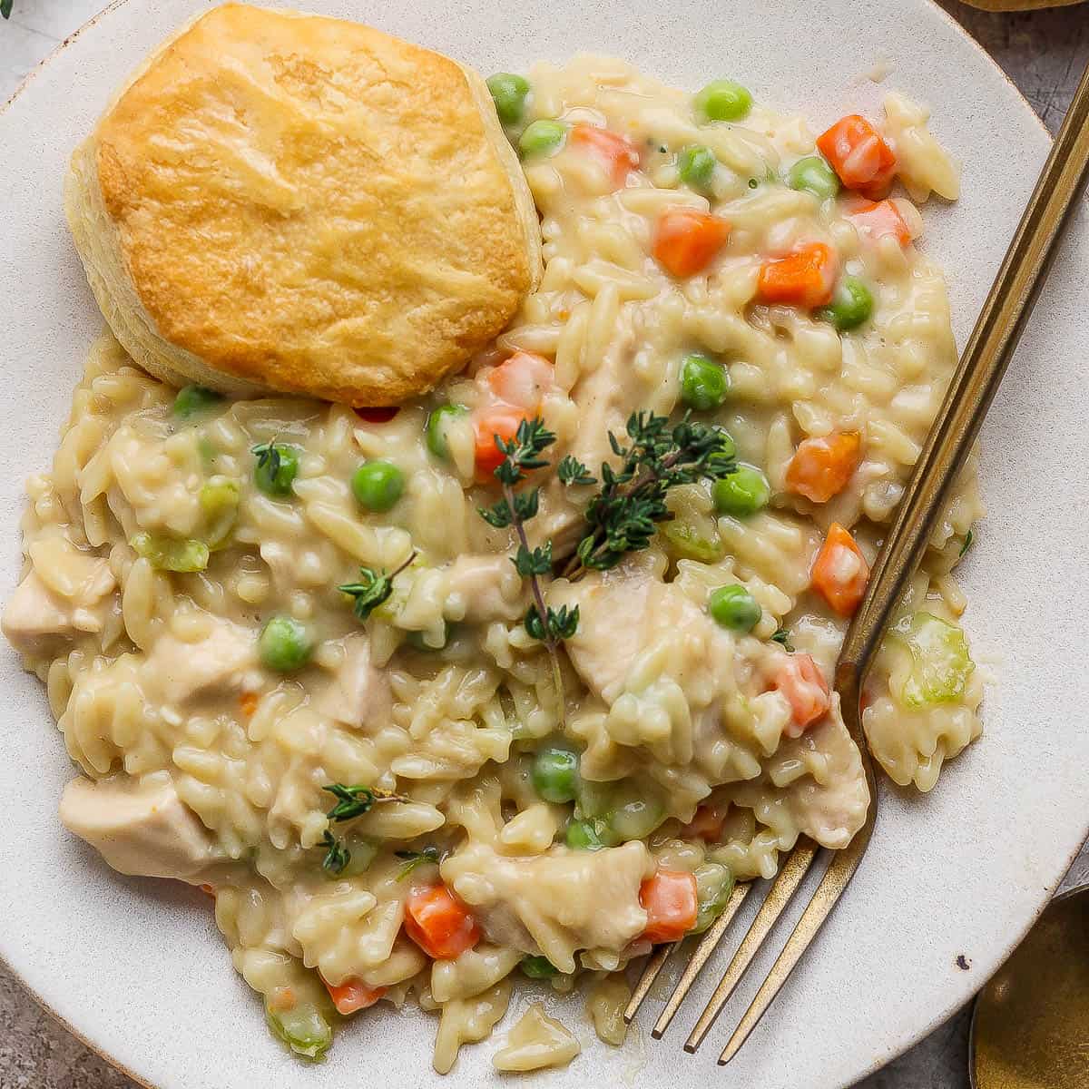 Chicken pot pie orzo on a white plate with a biscuit and gold fork.