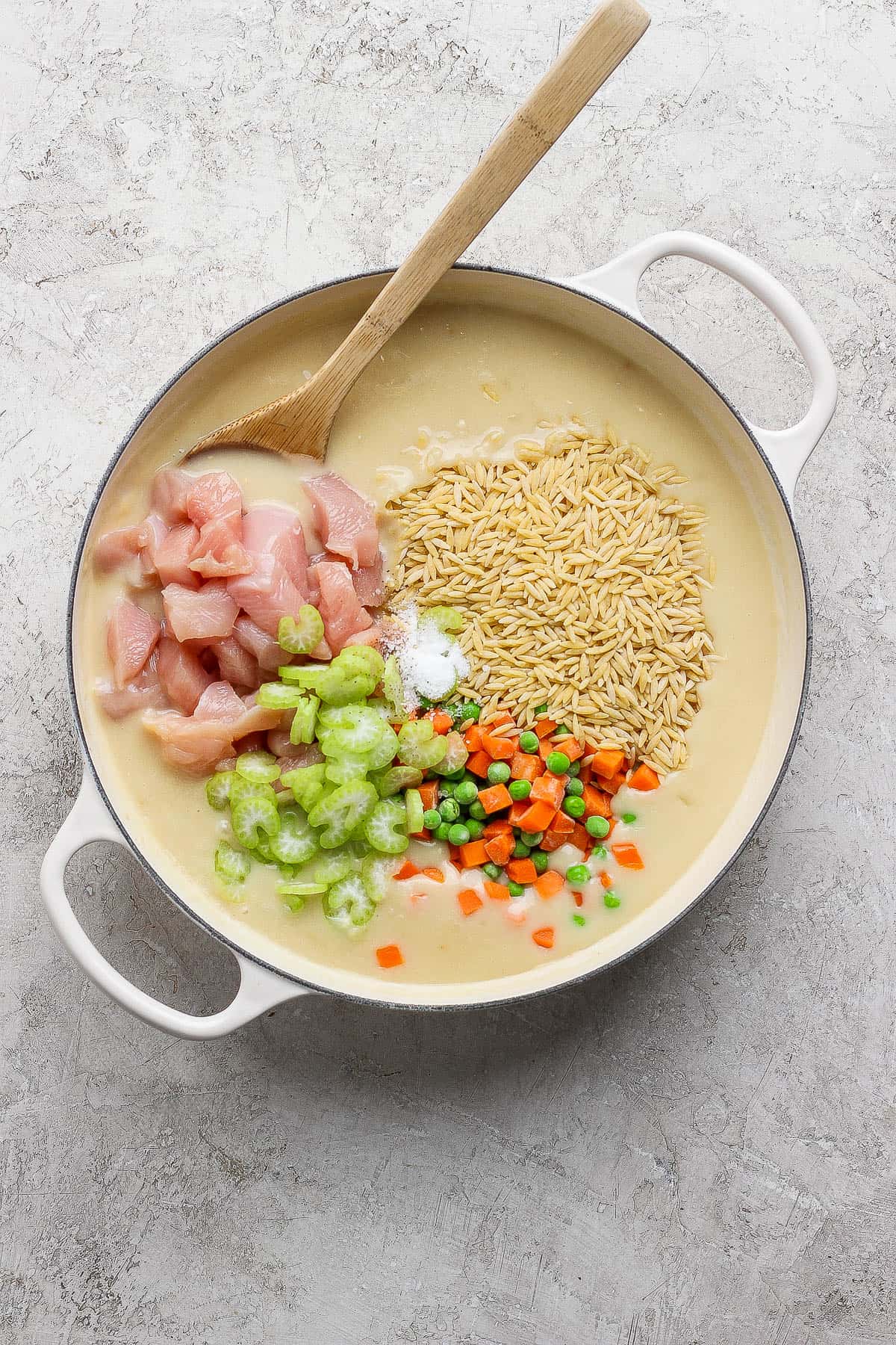 Raw chicken pieces, orzo, celery, frozen peas & carrots, and seasonings added to the creamy broth base of a one pan chicken pot pie orzo with a wooden spoon.