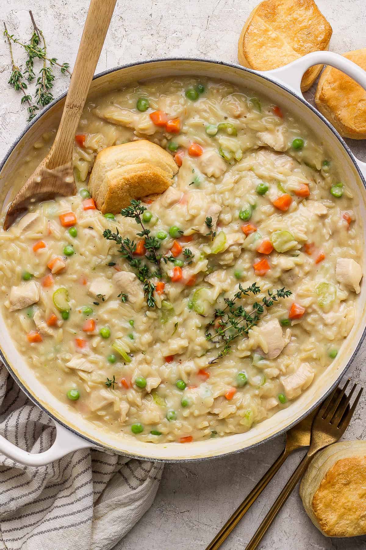 Chicken pot pie orzo in a braising pan with fresh thyme, a wooden spoon, and some biscuits.
