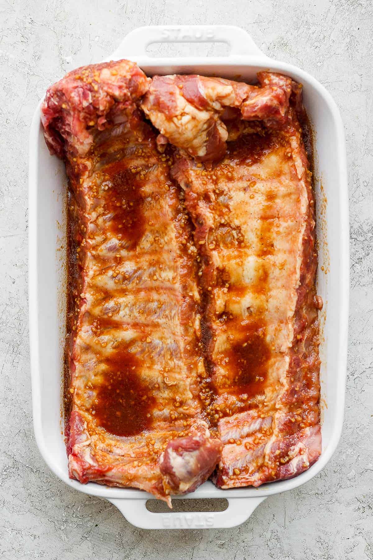 Two racks of baby back ribs marinating in a white baking dish.